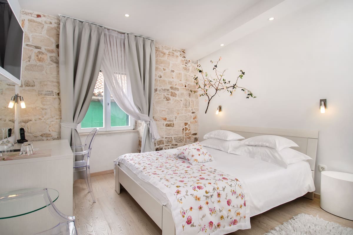 Un dormitorio con pared de piedra, una cama con ropa blanca, una mesa de vidrio y una ventana con cortinas.