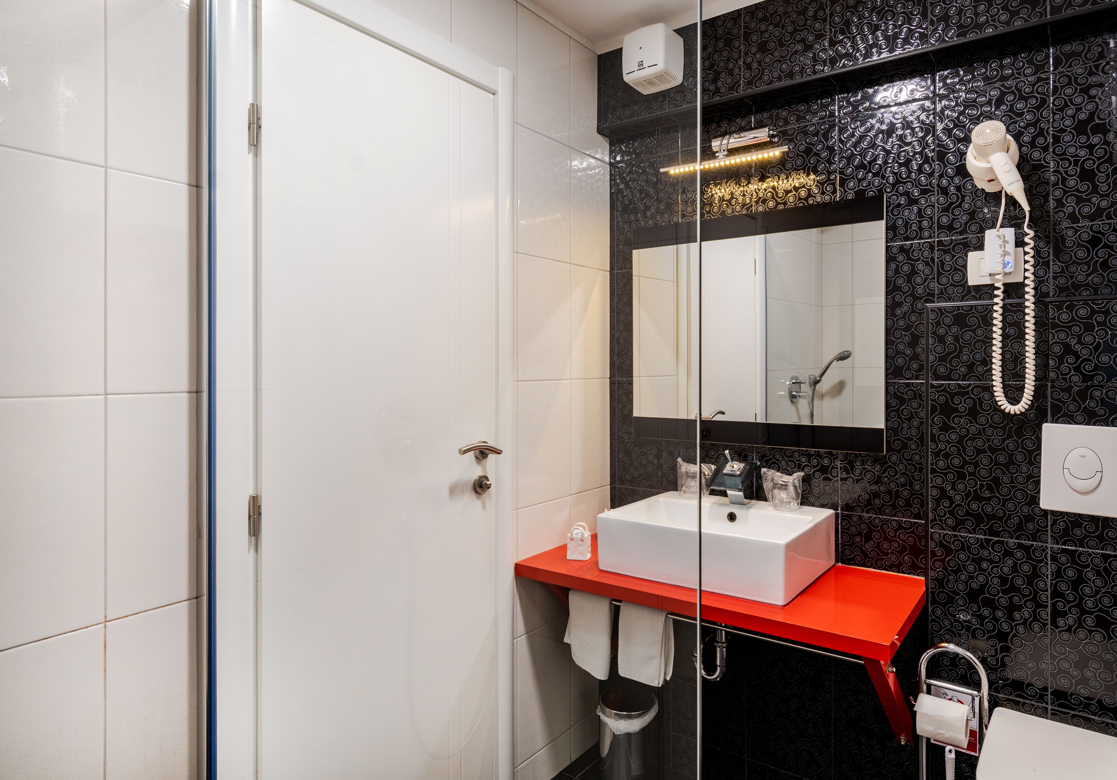 A modern bathroom with a white door, a red sink vanity, a glass shower enclosure, and a black tiled wall with a decorative pattern.