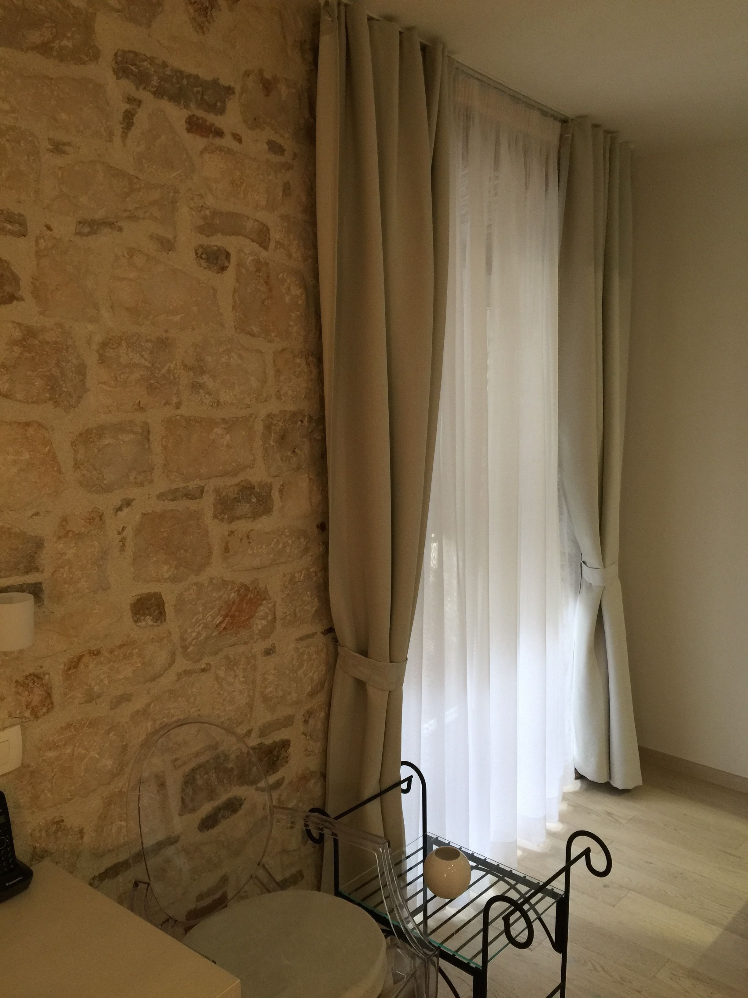 Una habitación con pared de piedra, una ventana con cortinas translúcidas, un perchero metálico con un jarrón y una silla blanca.