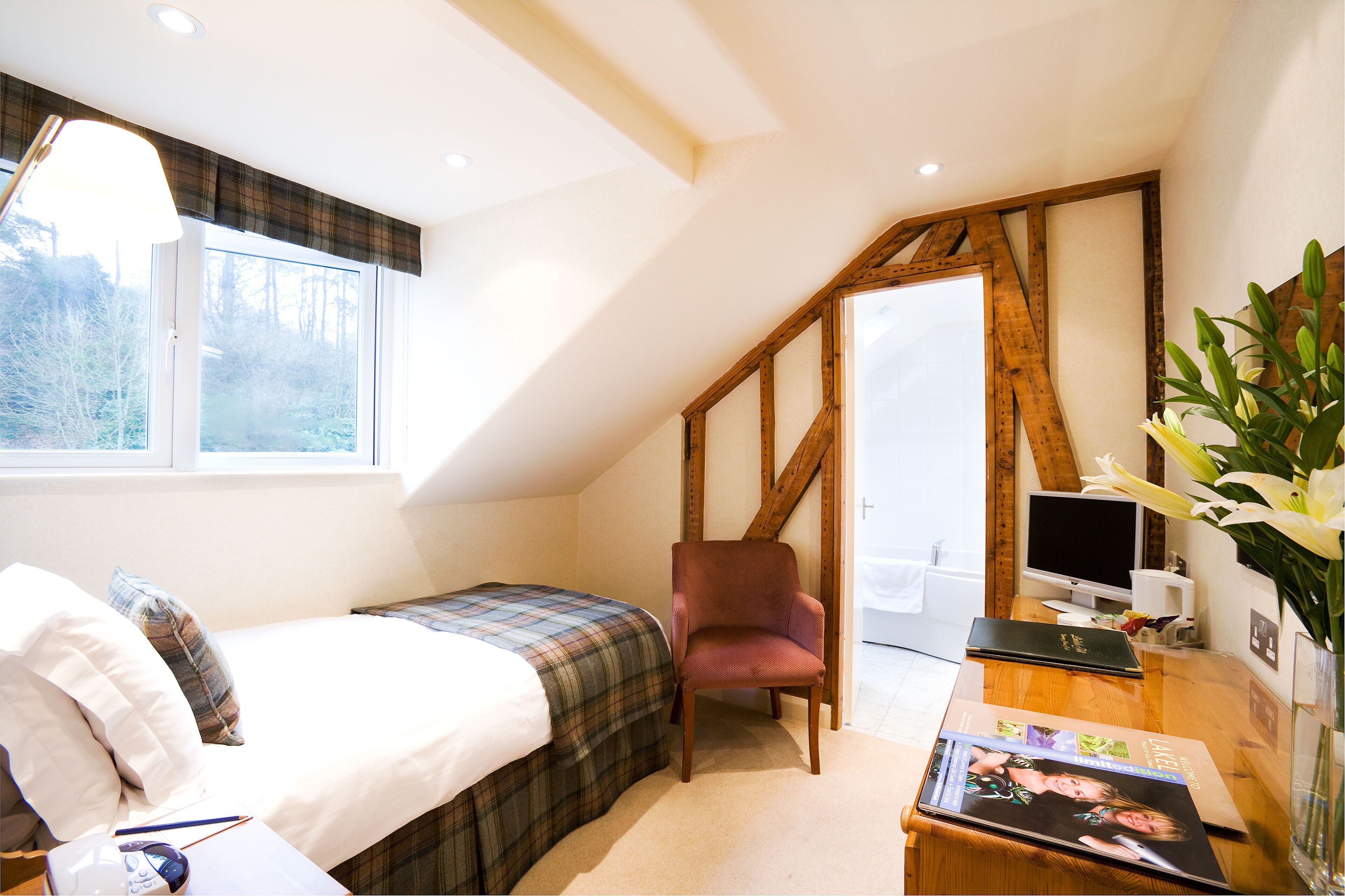 A bedroom with a bed, a chair, a desk with a computer, and a window with a view of trees.