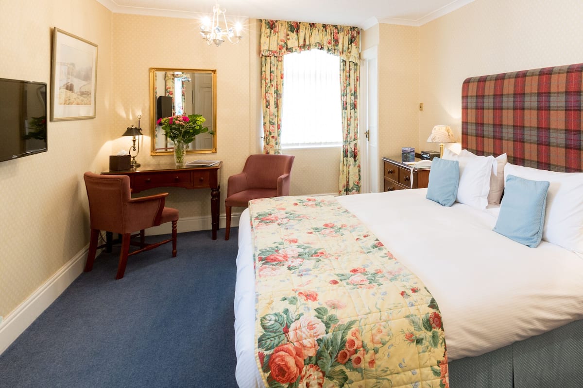 Une chambre d’hôtel avec un grand lit, un bureau avec une chaise, une commode, un miroir et une fenêtre avec des rideaux.