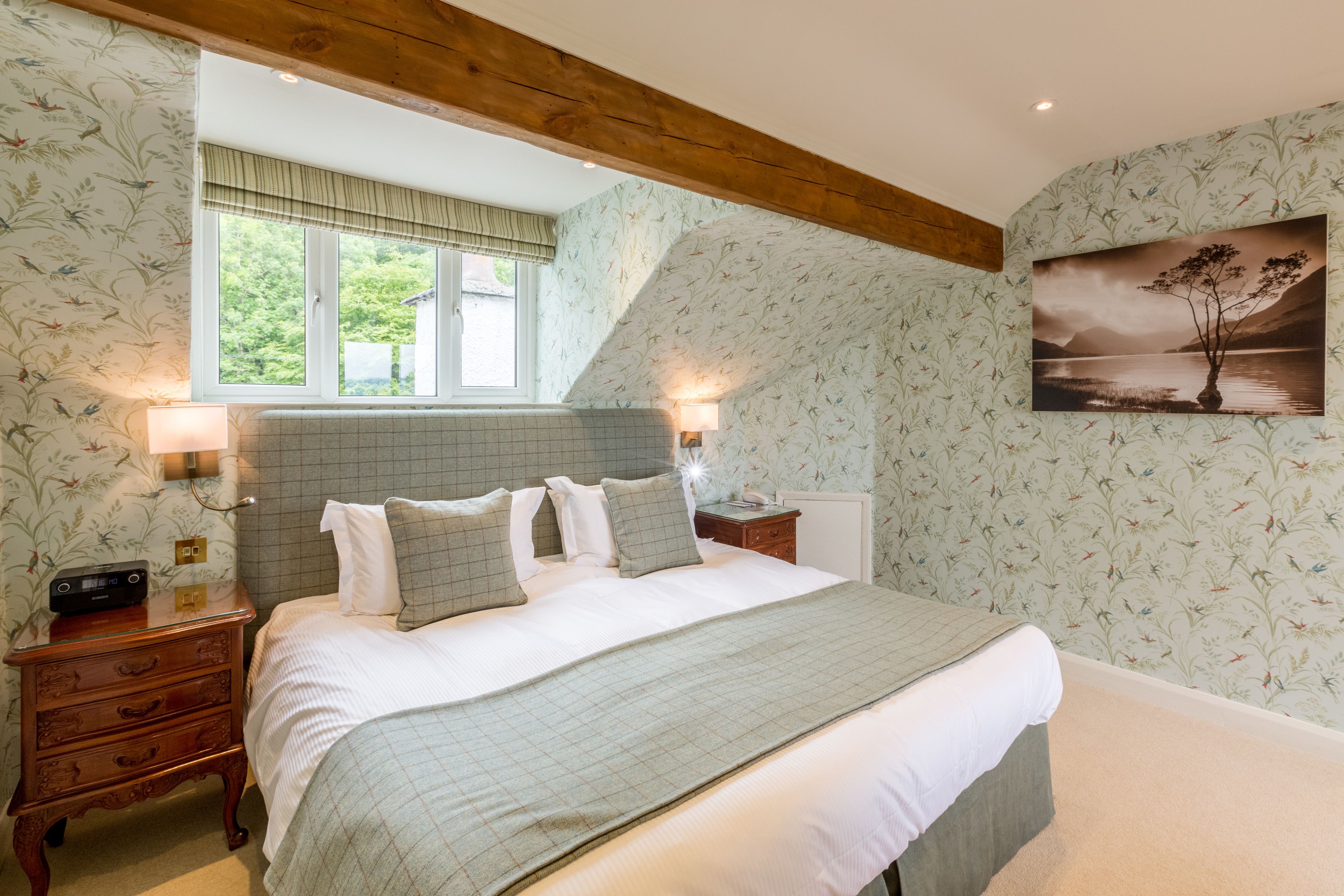A bedroom with a large bed, a nightstand, a lamp, a window with a view of greenery, and a wall with floral wallpaper.