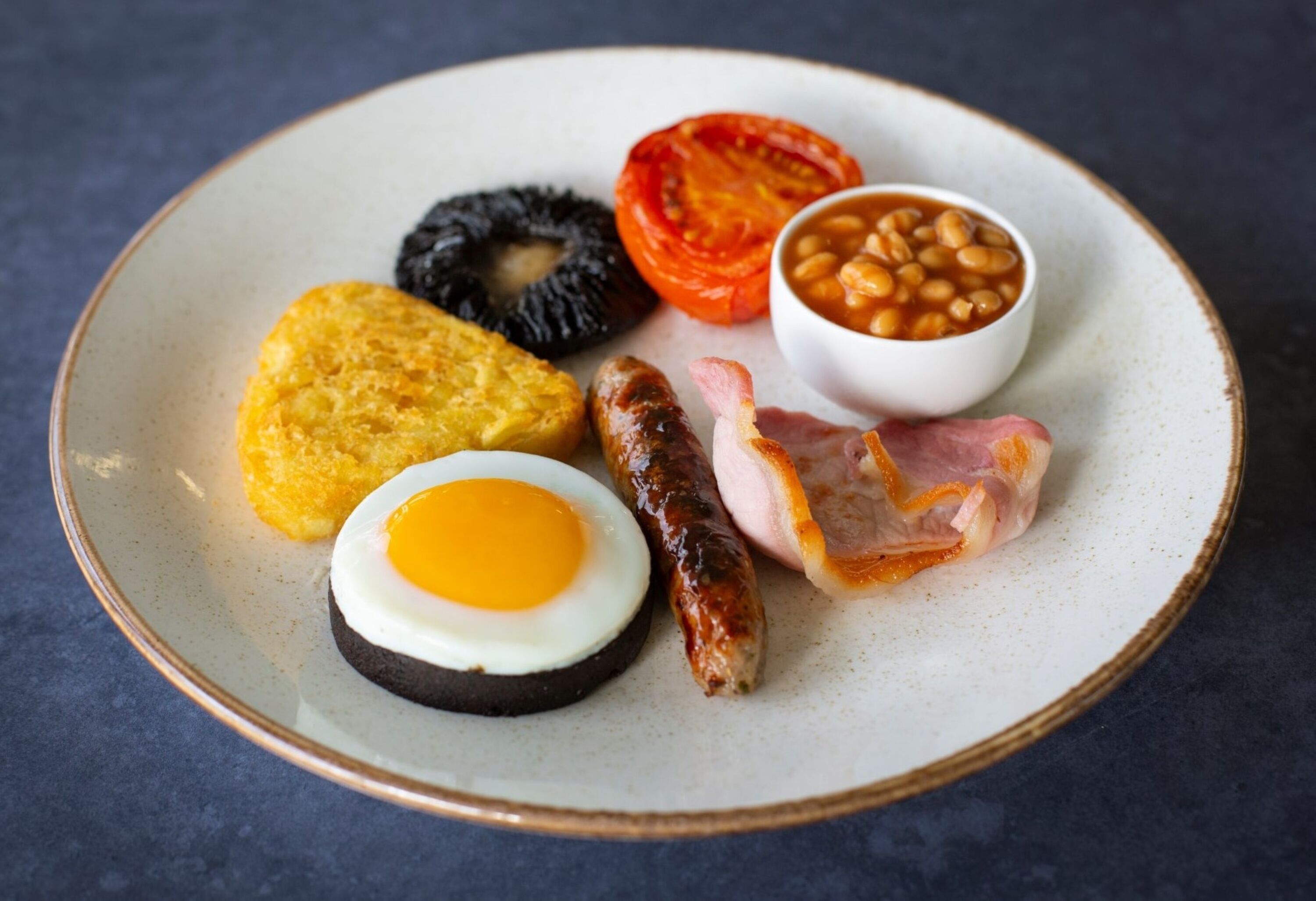 A plate with a variety of breakfast items including eggs, sausages, bacon, beans, a slice of tomato, and a piece of black pudding.