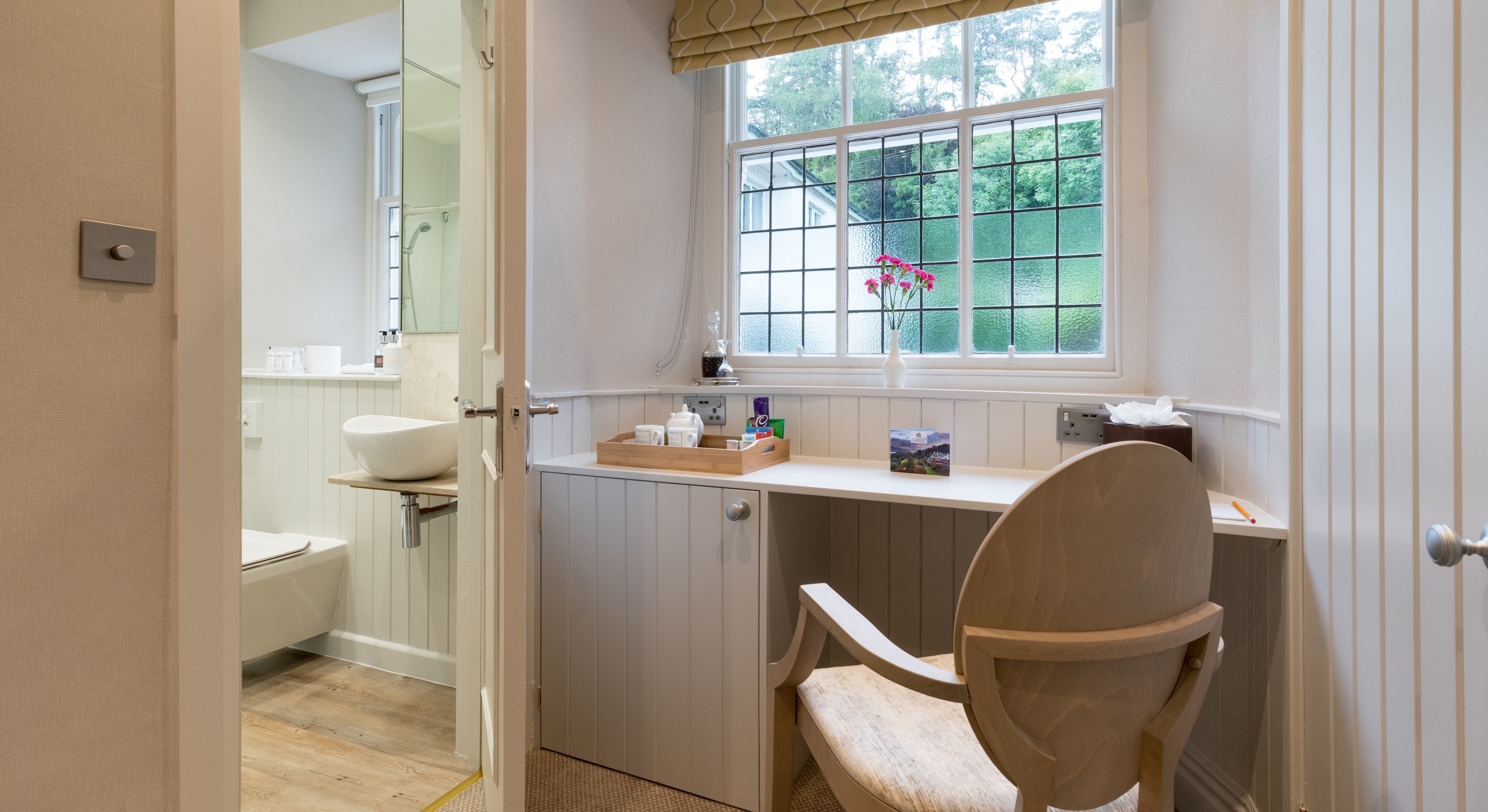 A bathroom with a white sink, a shower, a window with a view of greenery, and a wooden chair.
