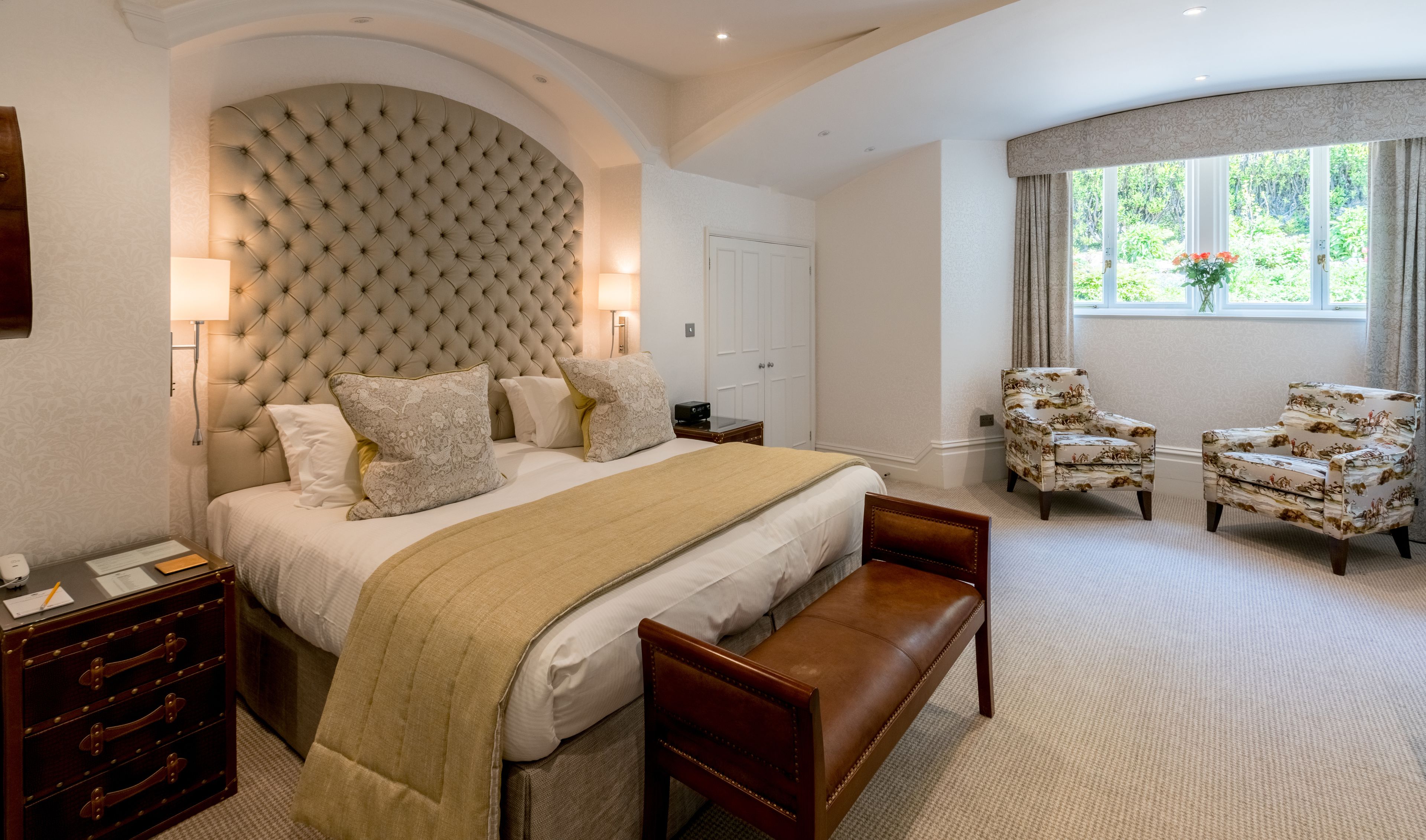 A bedroom with a large bed, a bench, two armchairs, a dresser, and a window with a view of greenery.