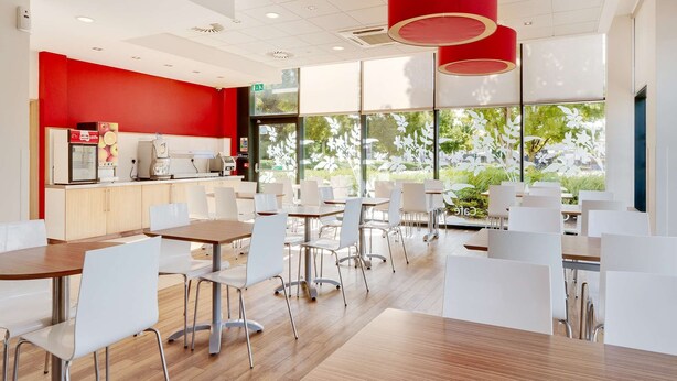 A modern cafe with white furniture, red accents, and large windows offering a view of greenery. - Travelodge Cardiff Atlantic Wharf Hotel (Cardiff)