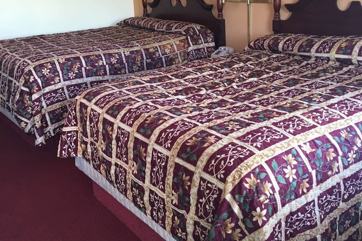 A hotel room with two beds, each with a floral-patterned bedspread, a nightstand with a lamp, and a wall-mounted smoke detector.