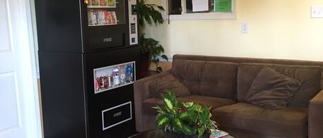 Un área de recepción con un refrigerador negro, un sofá marrón, una mesa de centro de vidrio con una planta y un piso de baldosas.