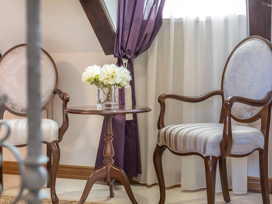 A room with two wooden chairs, a small round table with a vase of flowers, and a window with purple curtains.