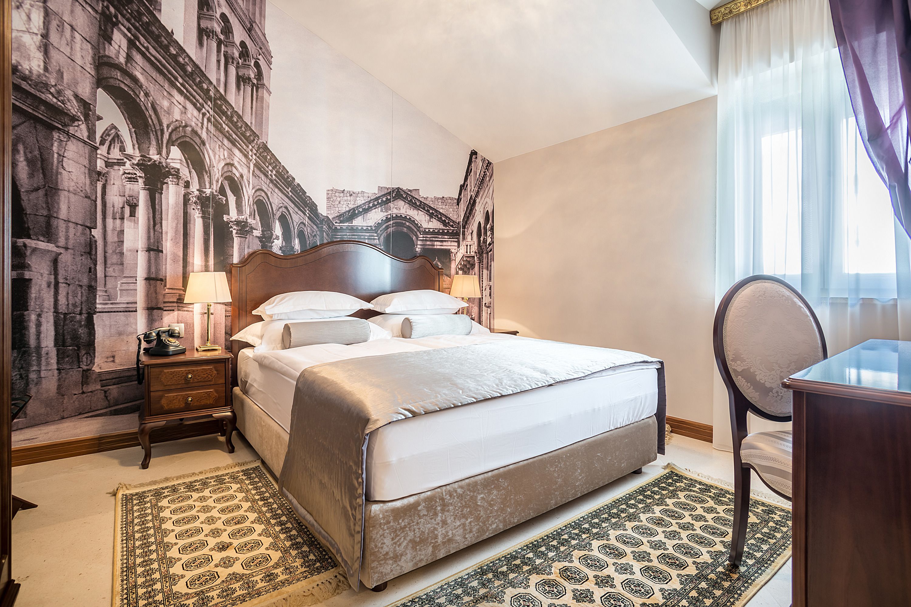 A hotel room with a large bed, a nightstand, a desk, and a chair. There is a large mural on the wall depicting an architectural scene.