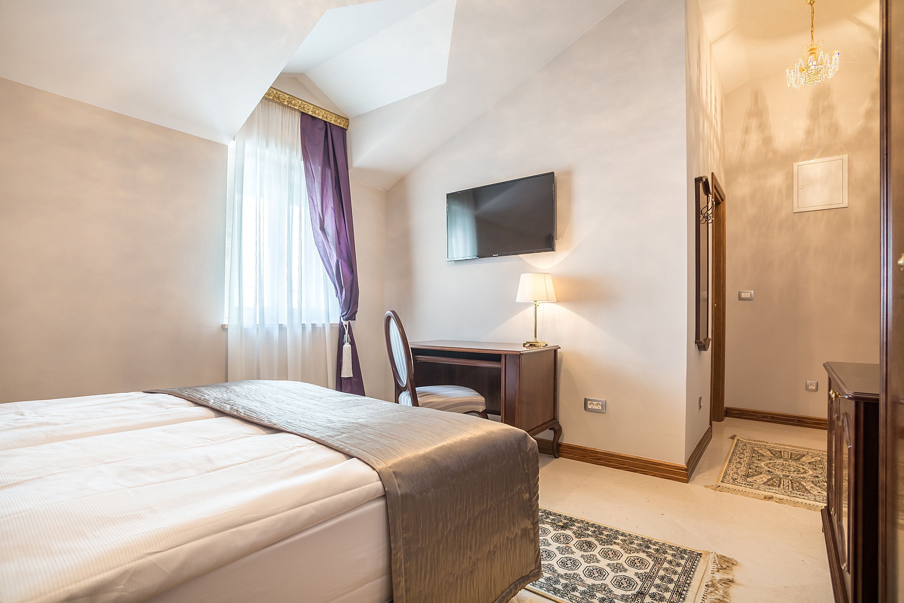 A hotel room with a large bed, a desk, a chair, a television, and a chandelier.
