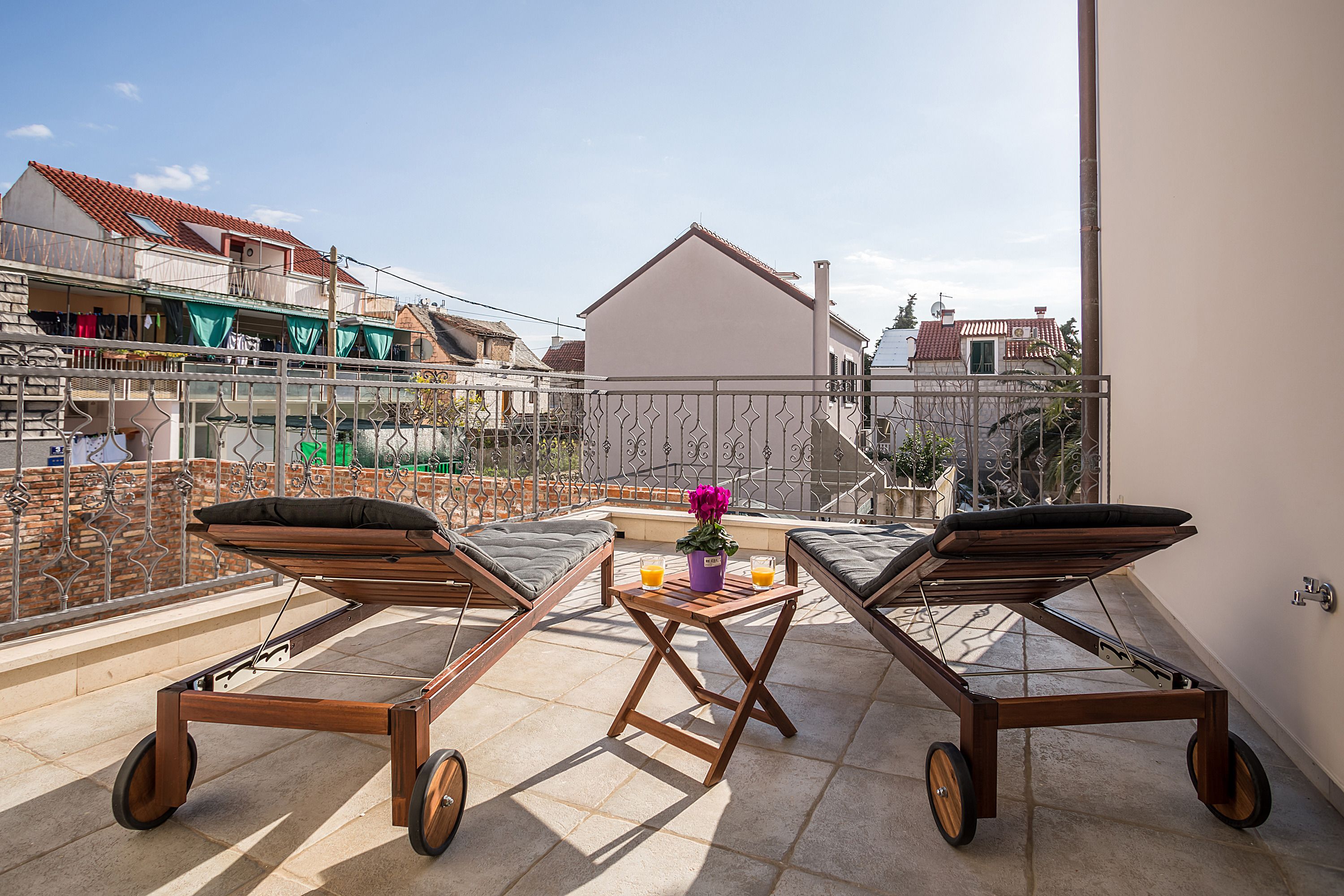 A balcony with two wooden loungers, a small table with a flower vase, and a view of neighboring buildings.