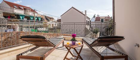 A balcony with two wooden loungers, a small table with a flower vase, and a view of neighboring buildings.