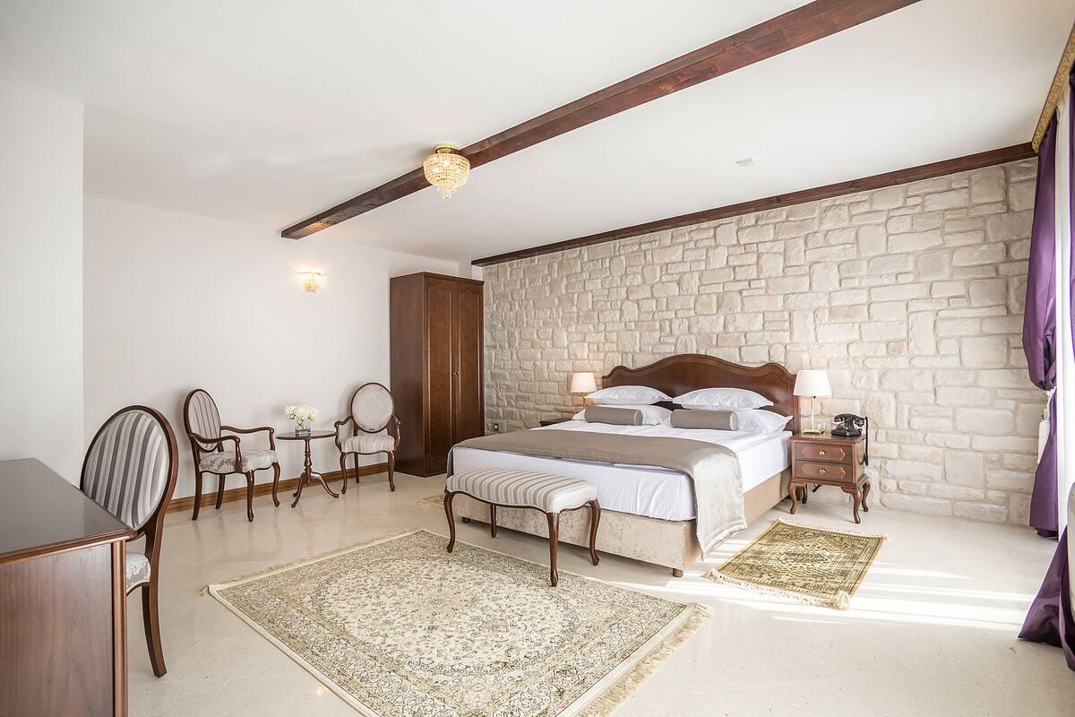 A bedroom with a stone wall, a wooden bed, a nightstand, a desk, and a chair.