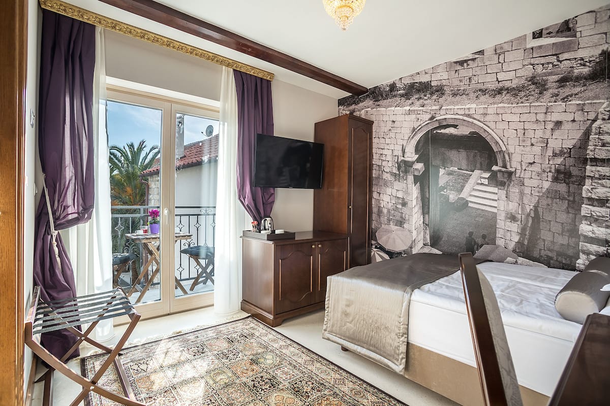 A bedroom with a bed, a wardrobe, a television, and a balcony with a view of palm trees.