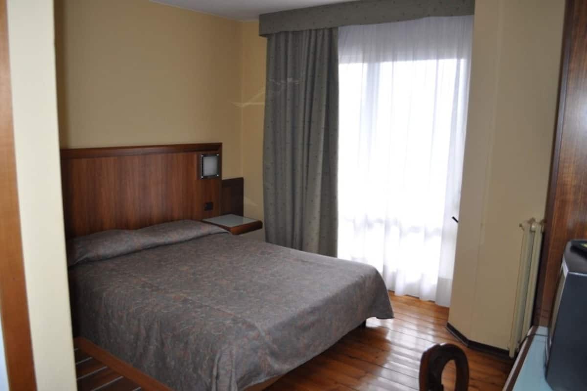 A hotel room with a single bed, a wooden headboard, a nightstand, a television, and a window with curtains.
