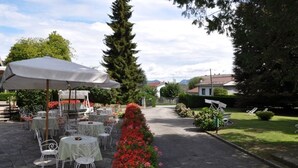 An outdoor patio with round tables and chairs, a large umbrella, and a flower bed. - Hotel Premeno (Premeno)