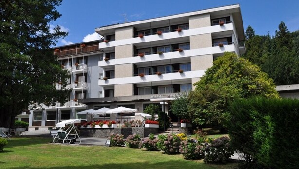 A multi-story hotel with a flat roof, balconies with flower boxes, and a well-maintained garden with outdoor seating. - Hotel Premeno (Premeno)