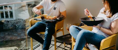 Duas pessoas sentadas em cadeiras de vime numa varanda, com uma mesa entre elas, uma taça de vinho e uma tigela de comida.