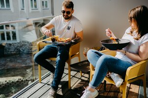 Duas pessoas sentadas em cadeiras de vime numa varanda, com uma mesa entre elas, uma taça de vinho e uma tigela de comida.