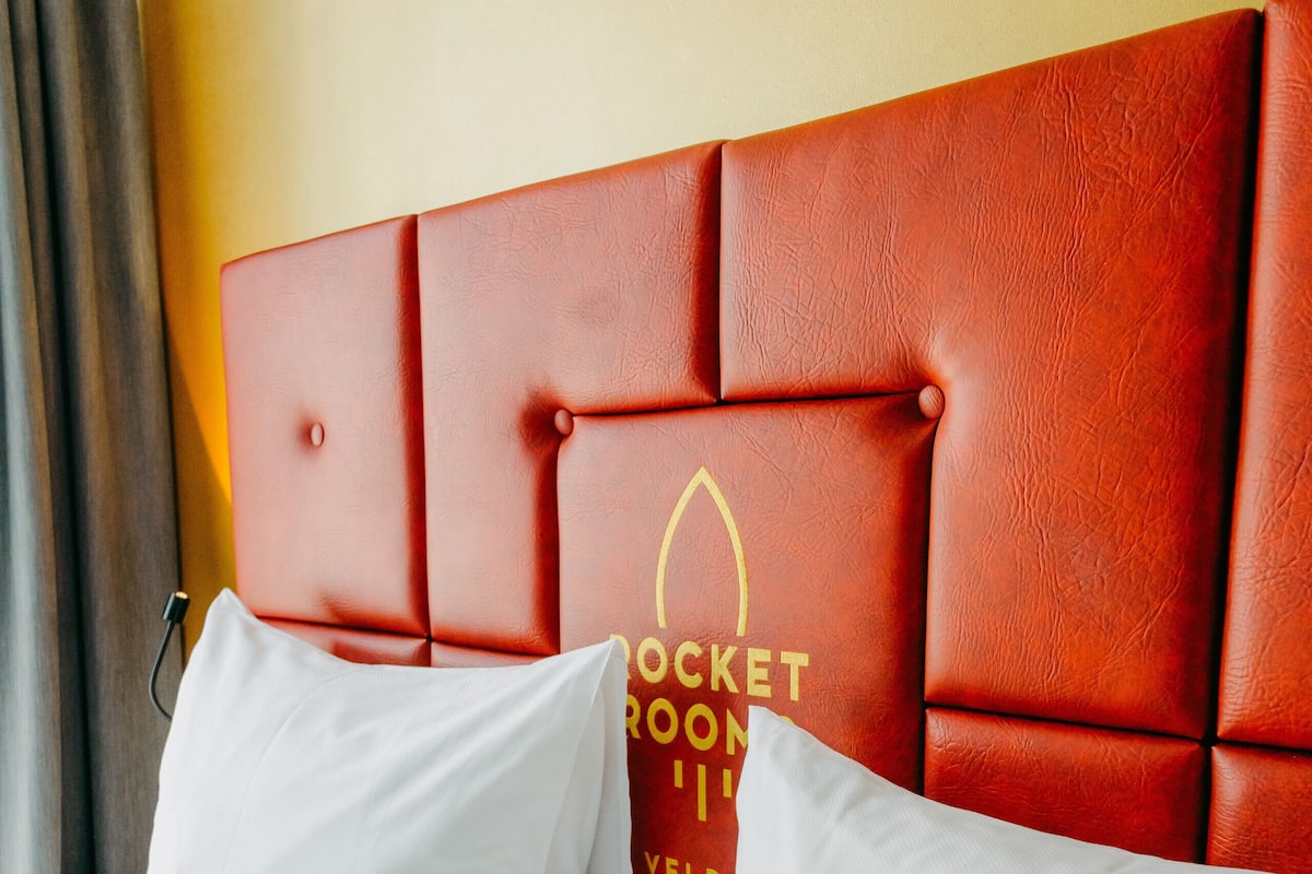 Ett hotellrum med en röd, klädd sänggavel, vita sängkläder och en logotyp på sänggaveln.