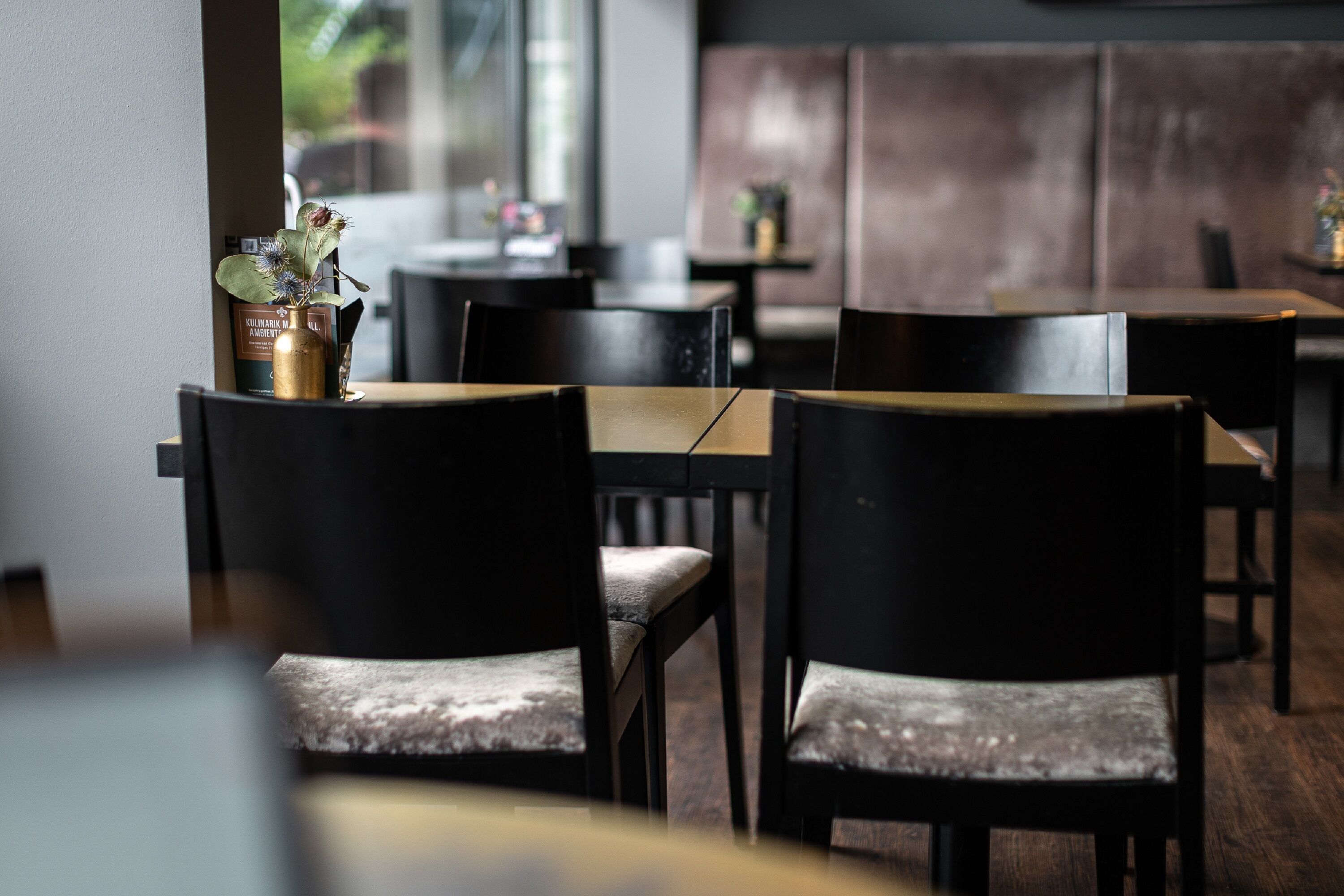 Un interior de restaurante con mesas y sillas de madera, un jarrón con flores y una ventana al fondo.