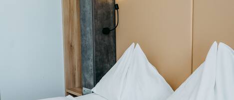 A bed with white linens, a wooden shelf, and a wall-mounted light fixture.