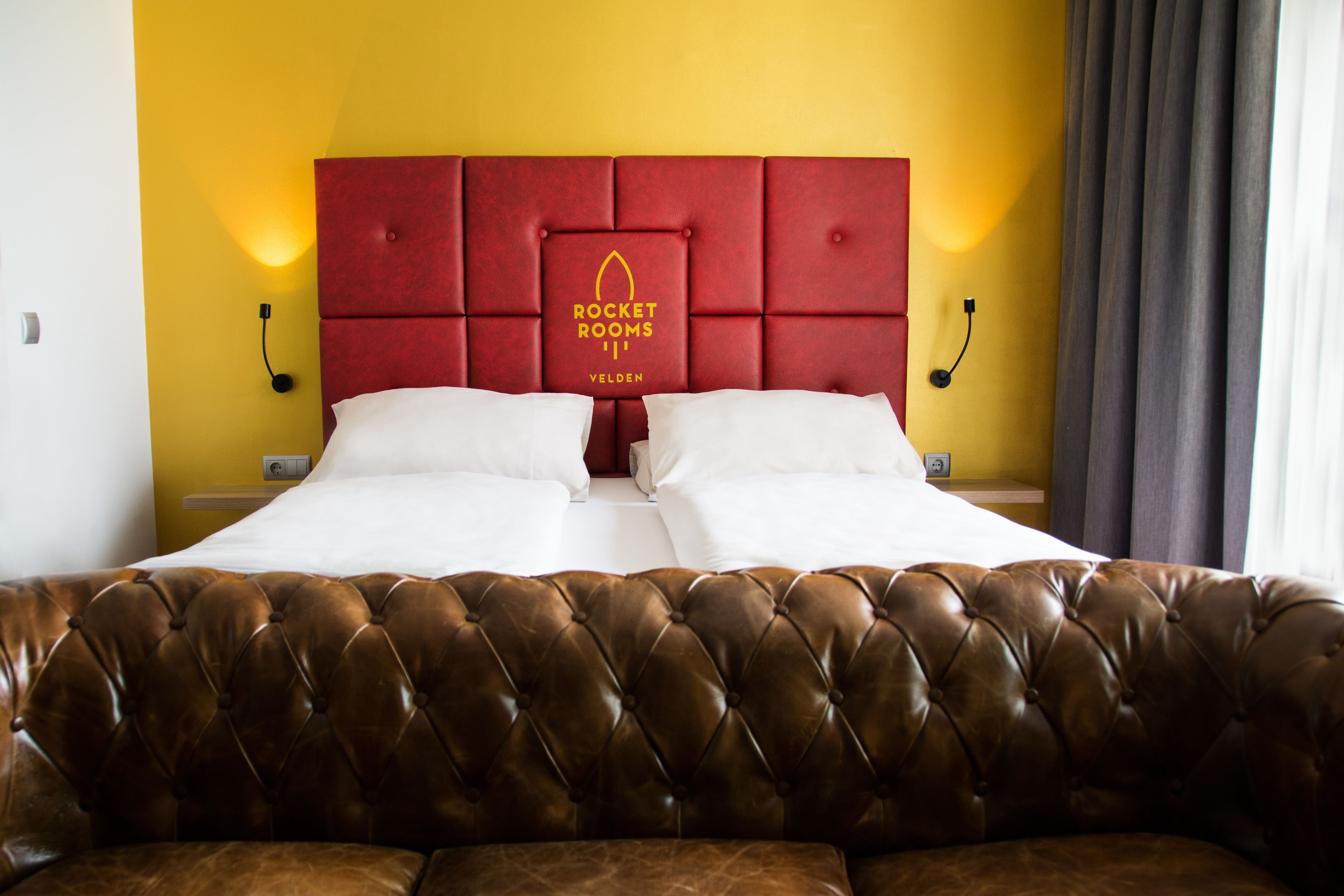 A hotel room with a red headboard, white bedding, and a brown leather sofa.