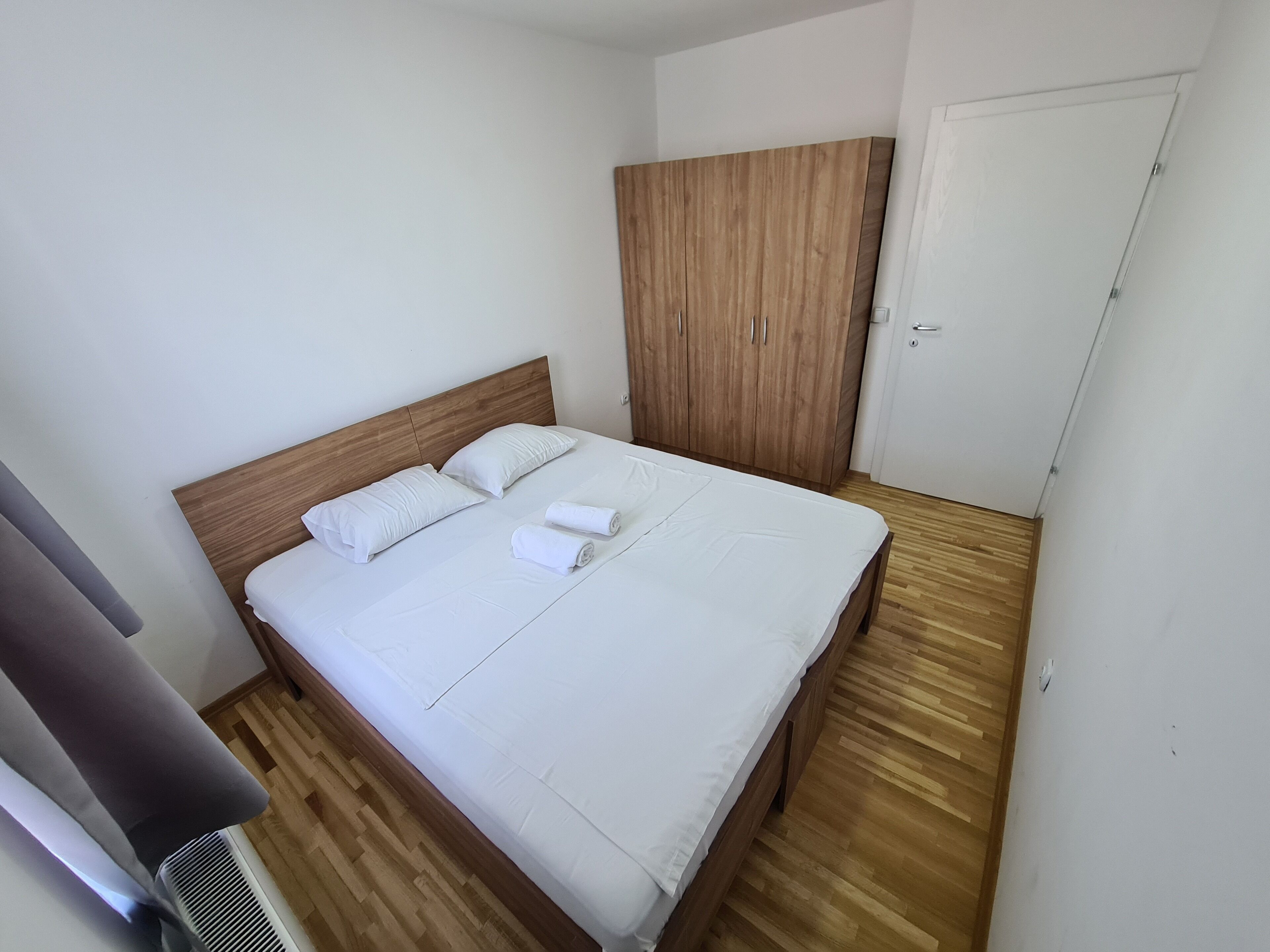A neatly made bed with white linens and two pillows, a wooden wardrobe, and a white door in a simple room with wooden flooring.