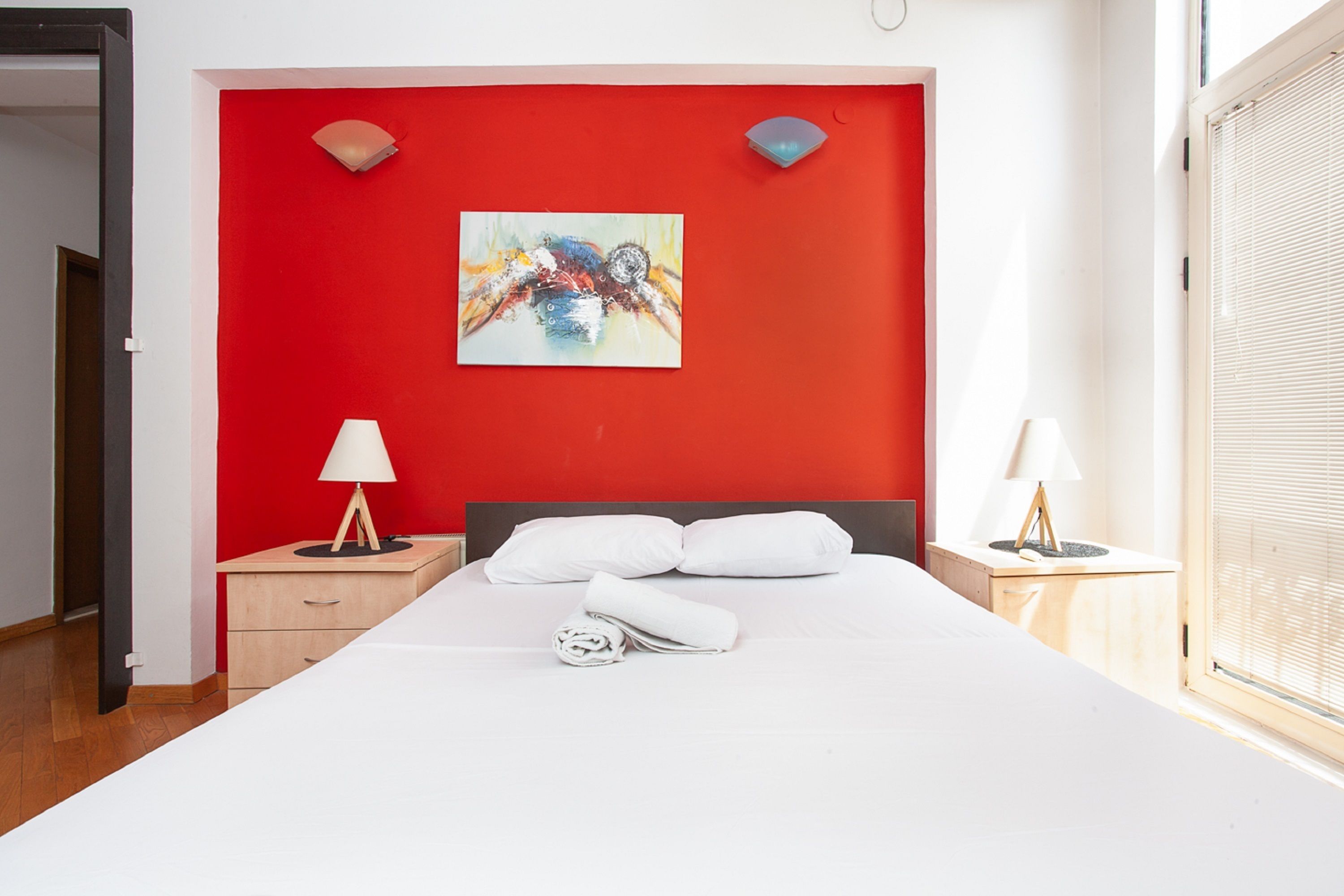 A hotel room with a red accent wall, a bed with white linens, two bedside tables with lamps, and a window with blinds.