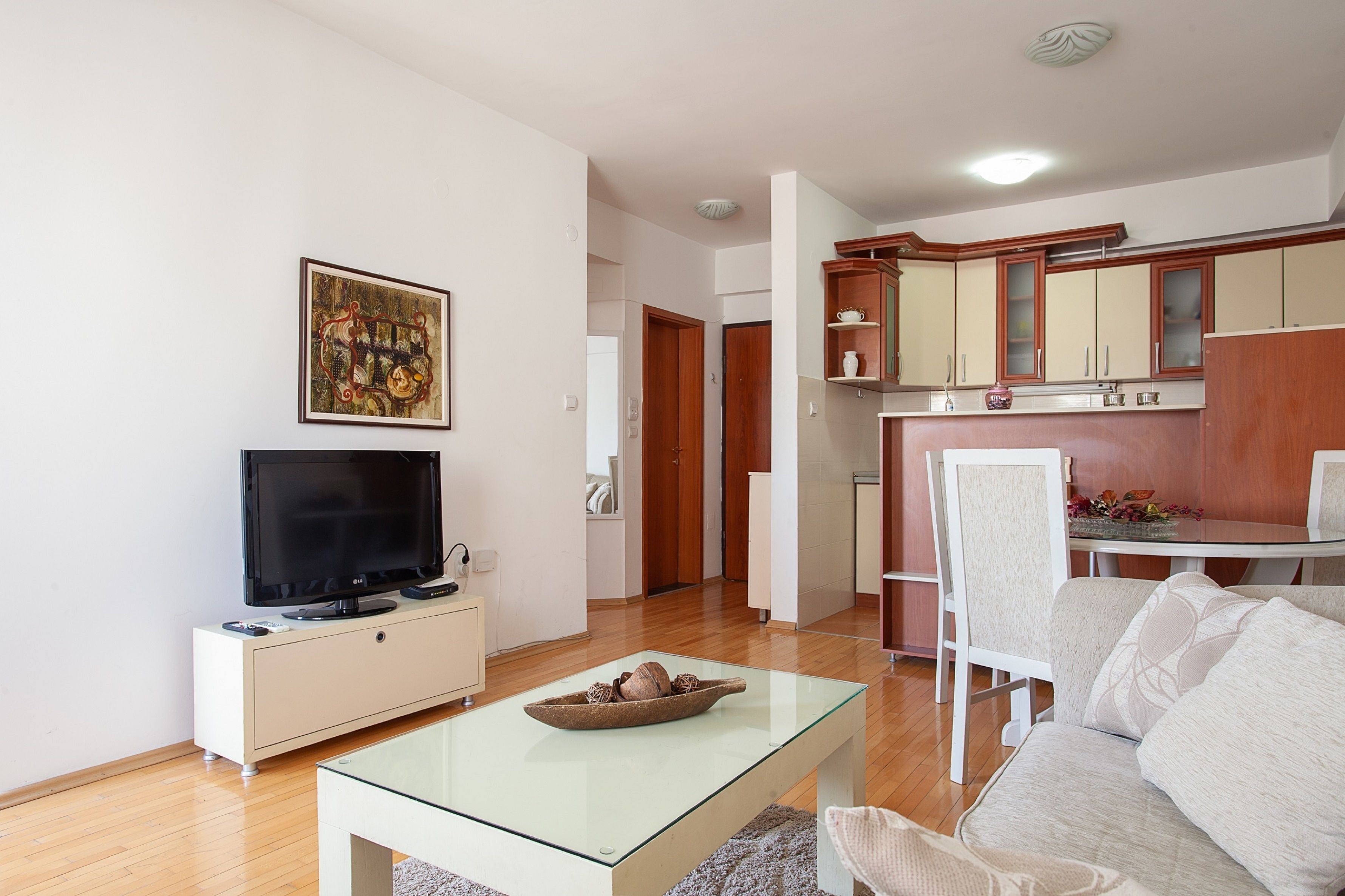 Una sala de estar con televisor, una mesa de centro de vidrio y un sofá. Hay una zona de cocina con armarios de madera y una mesa de comedor.