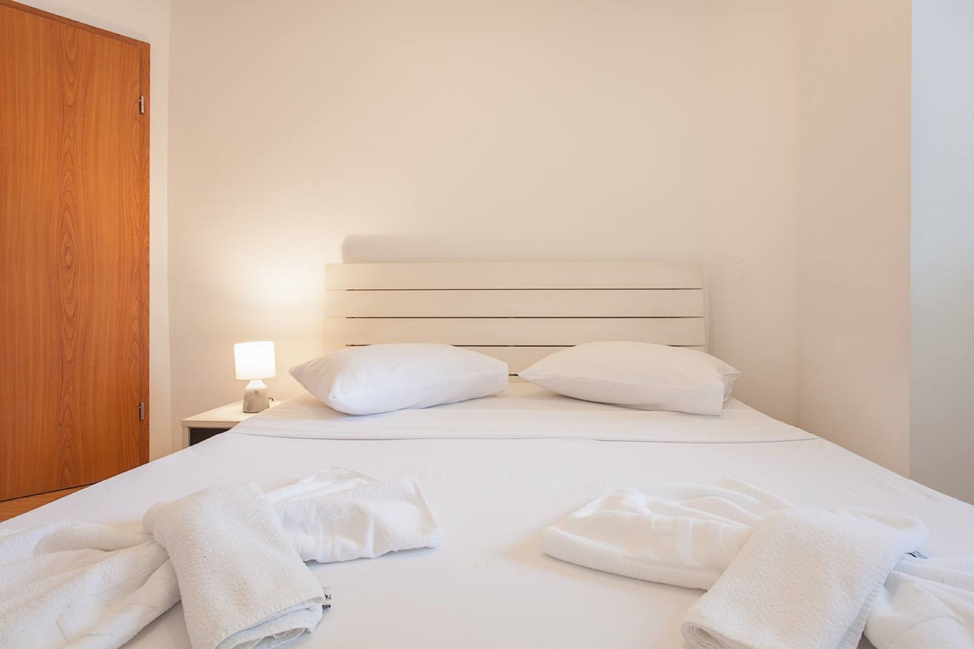 A neatly made bed with white linens and pillows, a wooden wardrobe, and a bedside lamp.