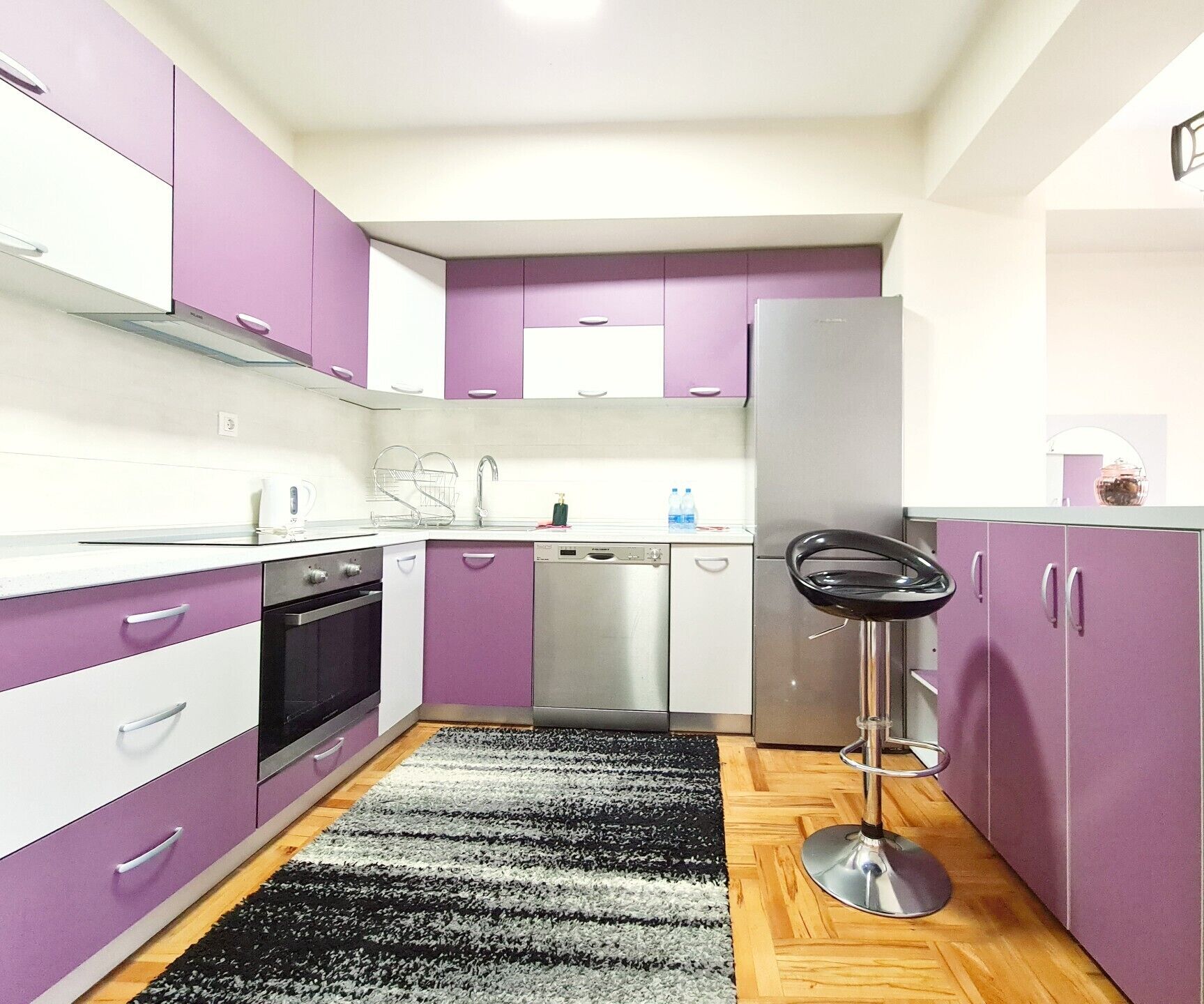 Una cocina compacta con armarios morados, un refrigerador de acero inoxidable y un taburete negro.
