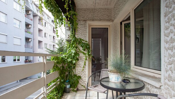 A balcony with a round table and chairs, a potted plant, and a view of a building with multiple windows and balconies. - Urban Hostel & Apartments (Skopje)