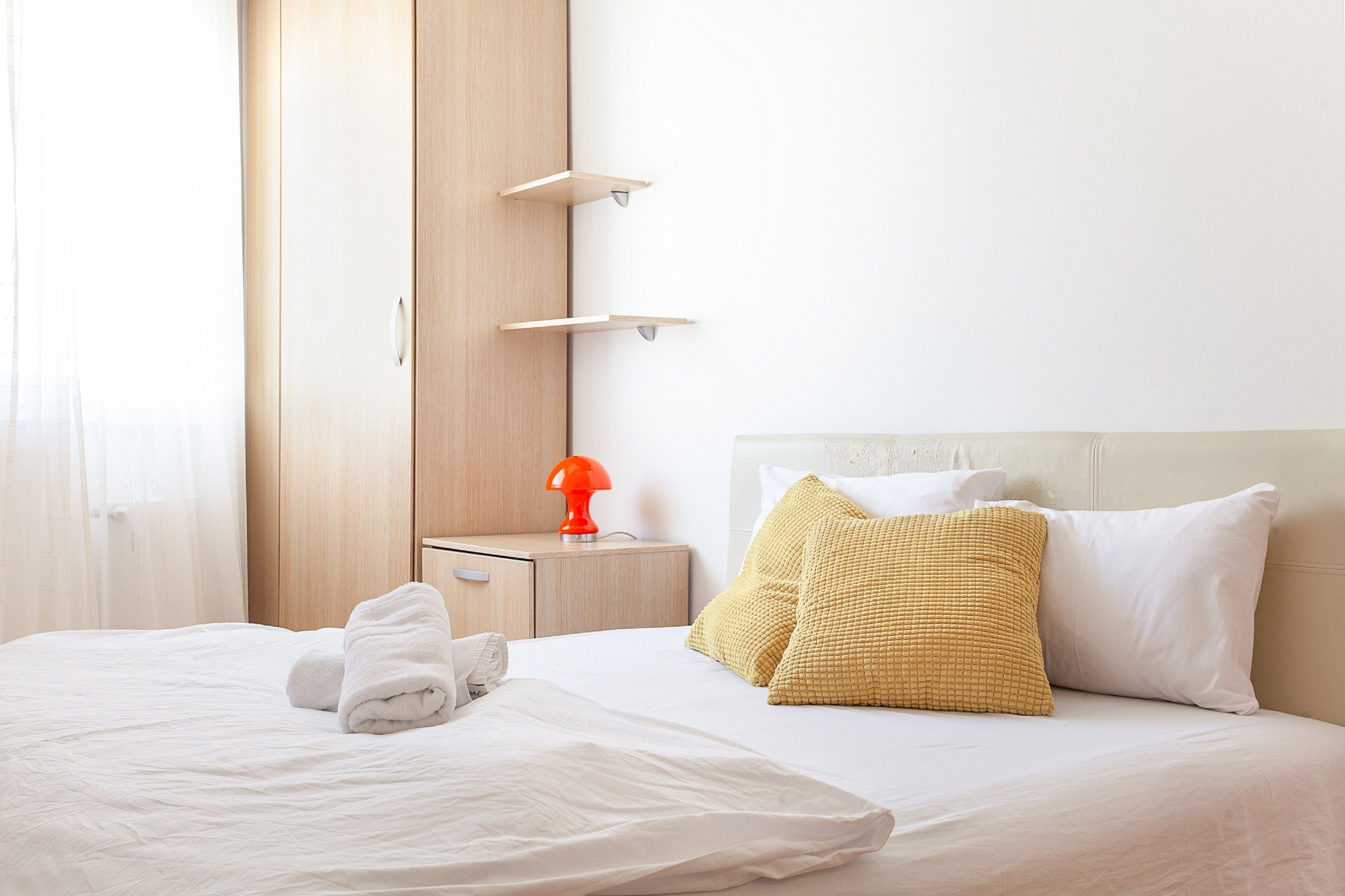 Cama bem arrumada com lençóis brancos, um travesseiro amarelo texturizado e um travesseiro branco. Há um guarda-roupa de madeira com porta e uma pequena mesa de cabeceira com um abajur vermelho.
