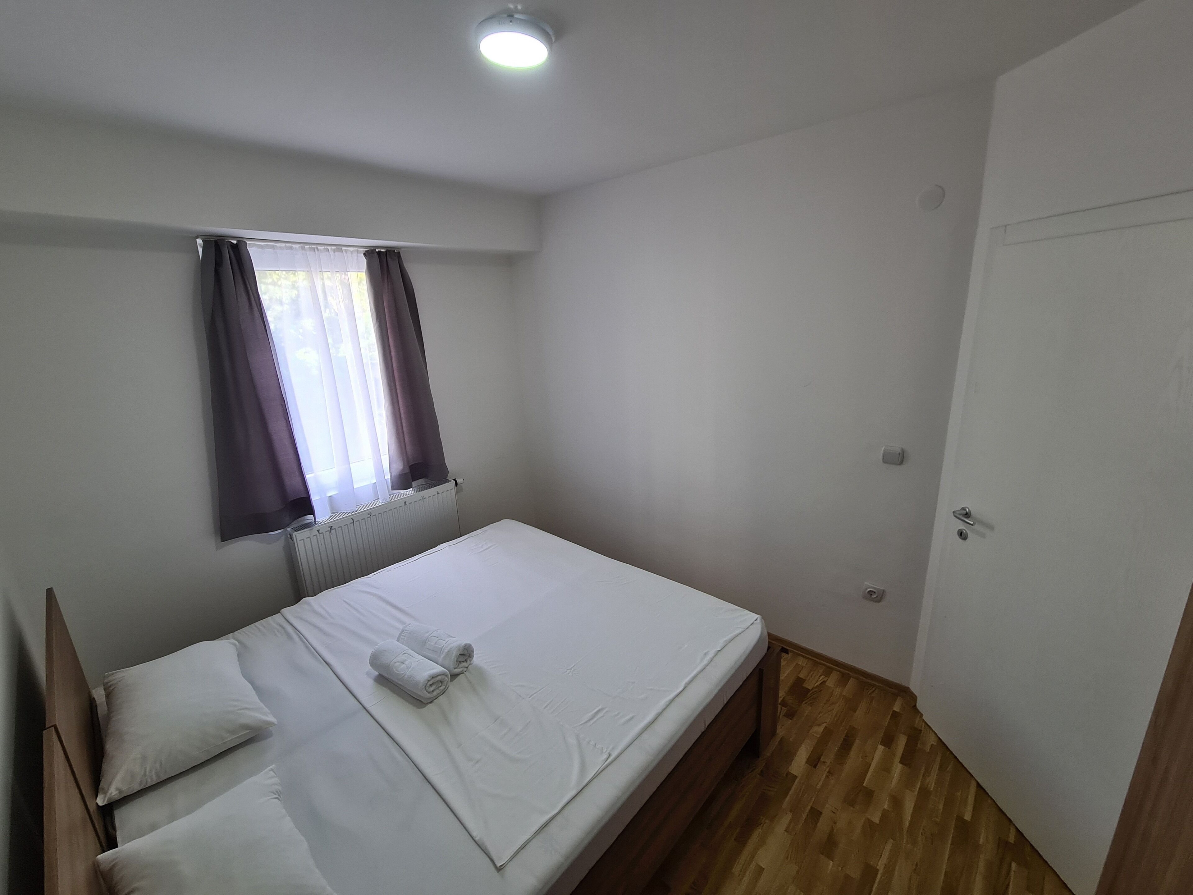 A single-bed room with a wooden floor, white walls, a window with curtains, and a door.