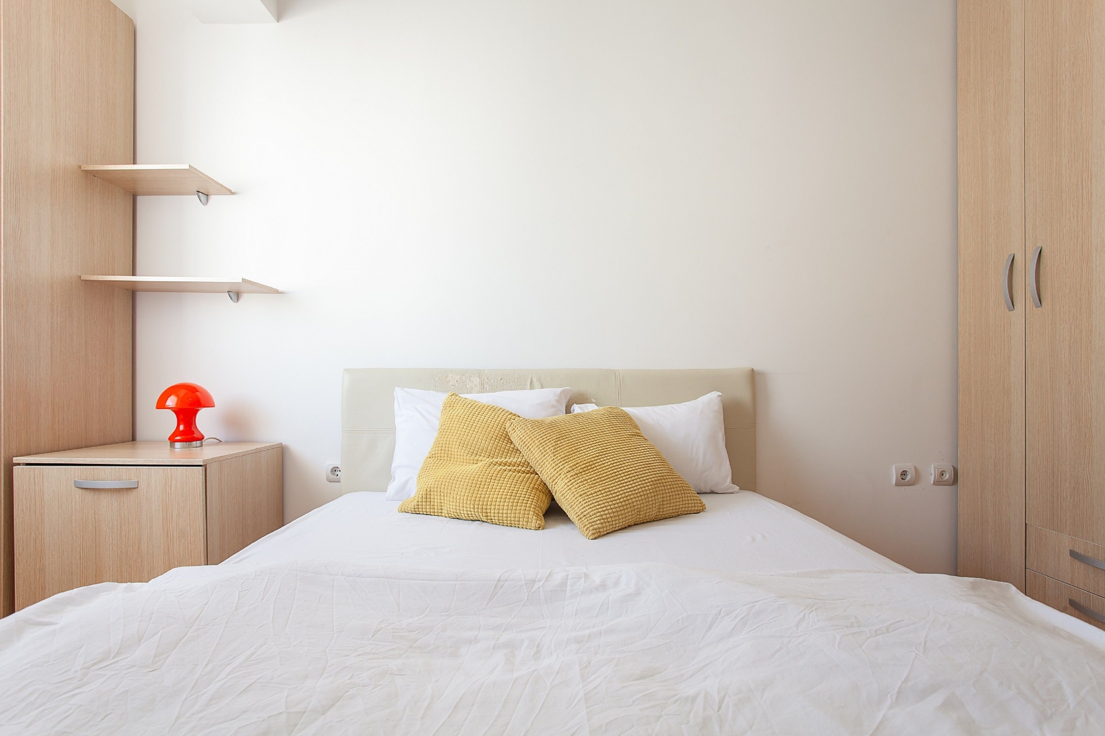 A neatly made bed with white bedding and two yellow pillows, a small bedside table with a red lamp, and a wooden wardrobe.