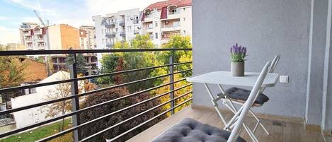 Un balcón con mesa y sillas, una planta en maceta y vistas a edificios y árboles.