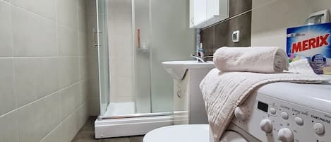 A compact bathroom with a glass shower enclosure, a toilet, and a washing machine.
