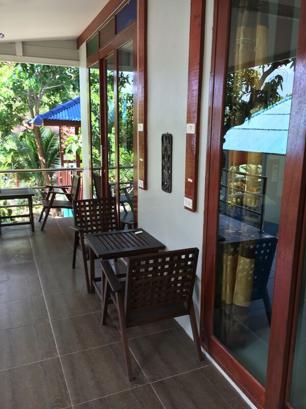 A covered outdoor seating area with wooden tables and chairs, a glass wall with a view of greenery, and a blue awning. - Reggae Village (Ko Pha-ngan)