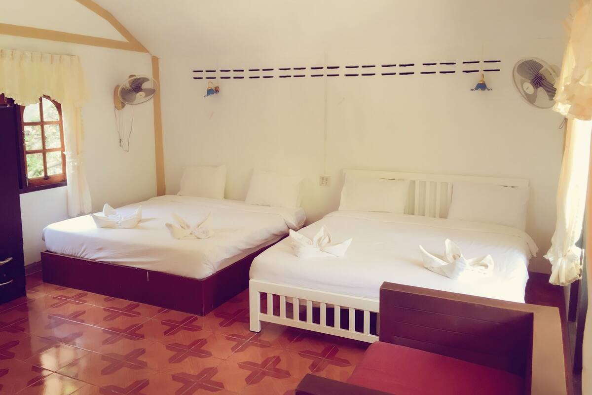 Ein Hotelzimmer mit zwei Betten, Holzboden, einem Fenster mit Vorhängen und zwei Wandventilatoren.