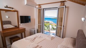 A hotel room with a bed, a desk, a mirror, a TV, and a balcony with a view of the sea. - Oasis Hotel (Lefkada)