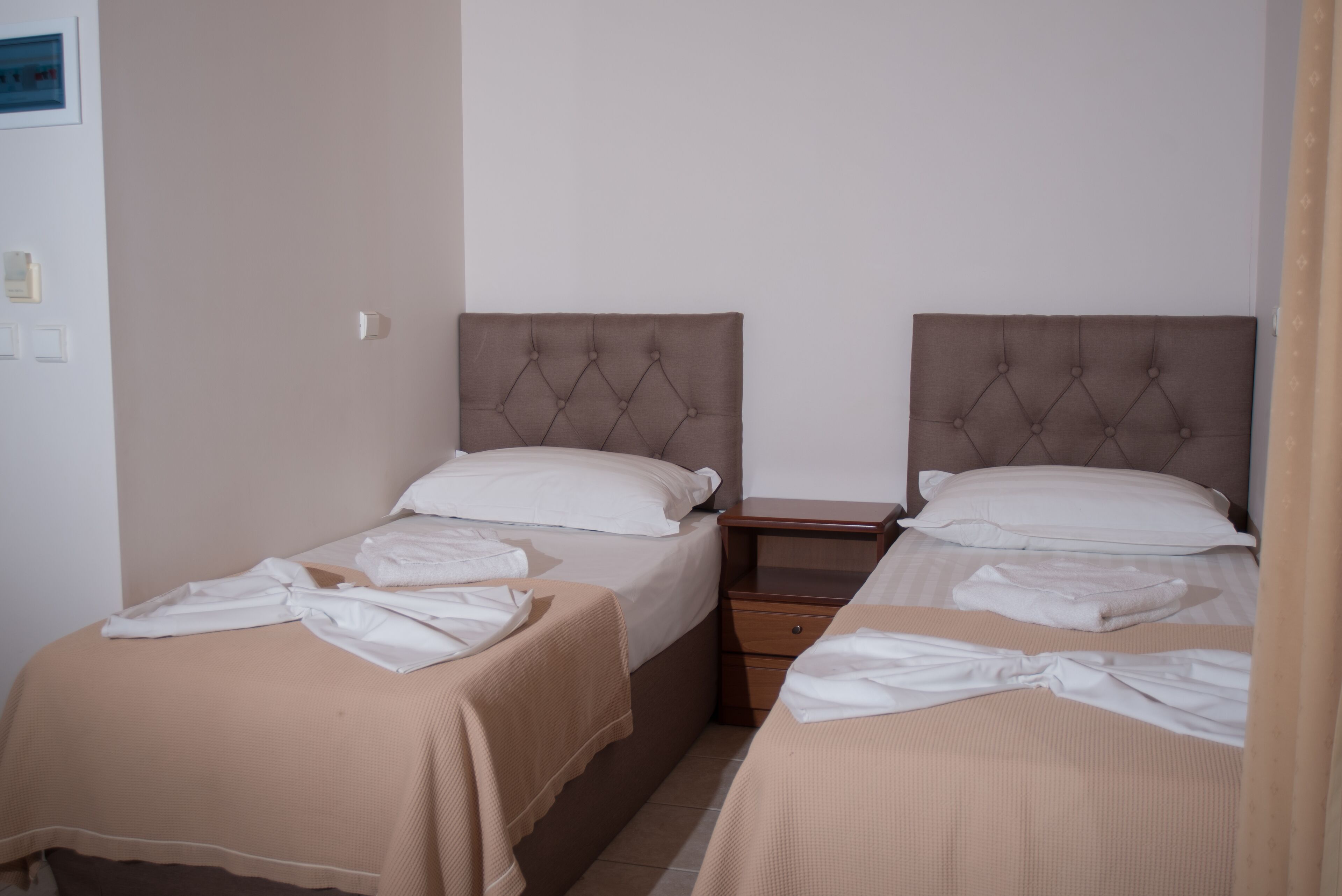 Habitación de hotel con dos camas, cada una con cabecero marrón y ropa de cama blanca. Hay una mesita de noche de madera entre las camas y una pequeña mesa con una lámpara.