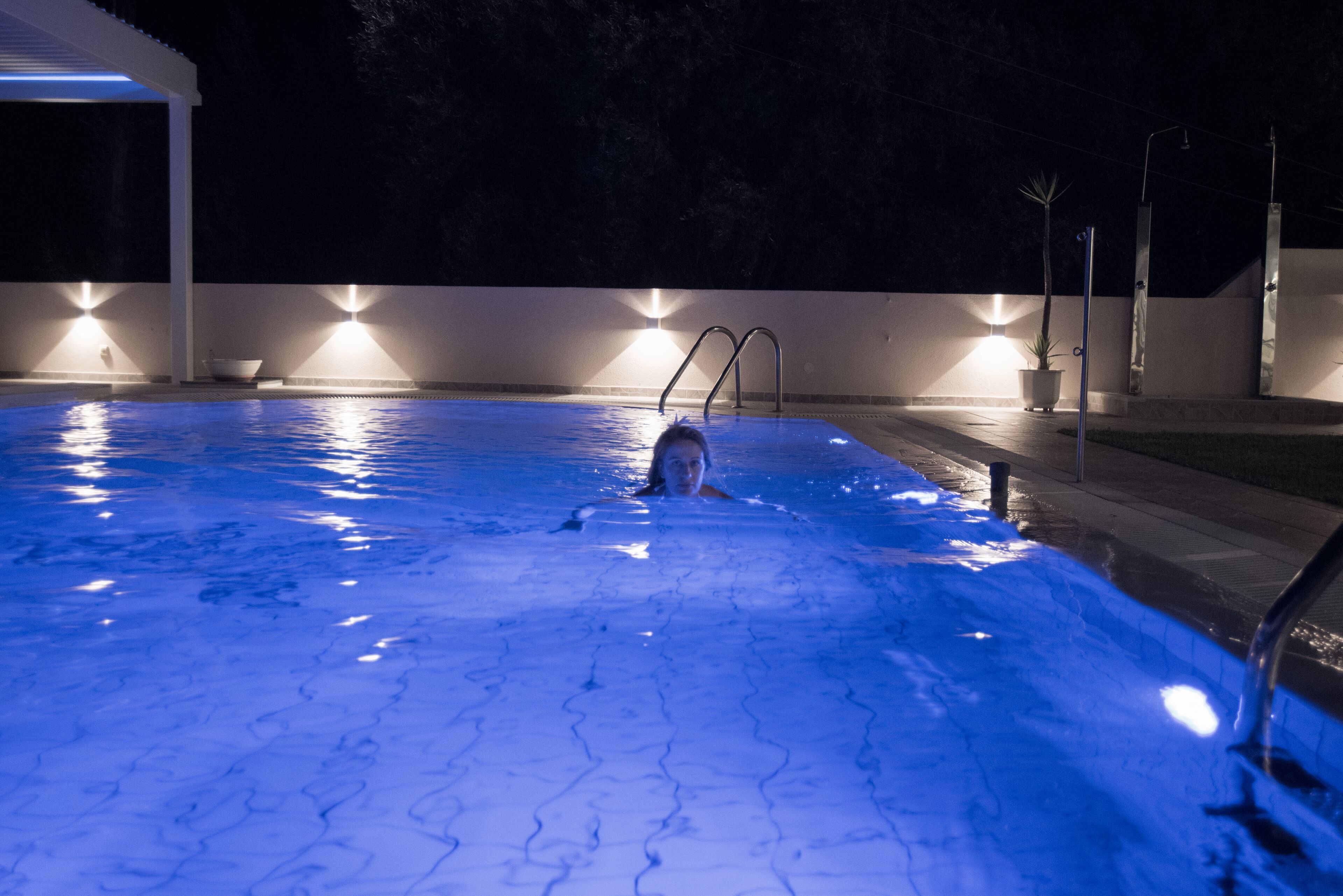 A swimming pool at night with a person swimming, surrounded by illuminated walls and a covered area.