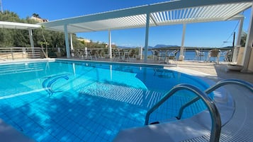 Ein klares, blaues Schwimmbecken mit Delfin-Motiv, umgeben von einer weißen Pergola und Stühlen, mit Blick auf ein Gewässer und Hügel.