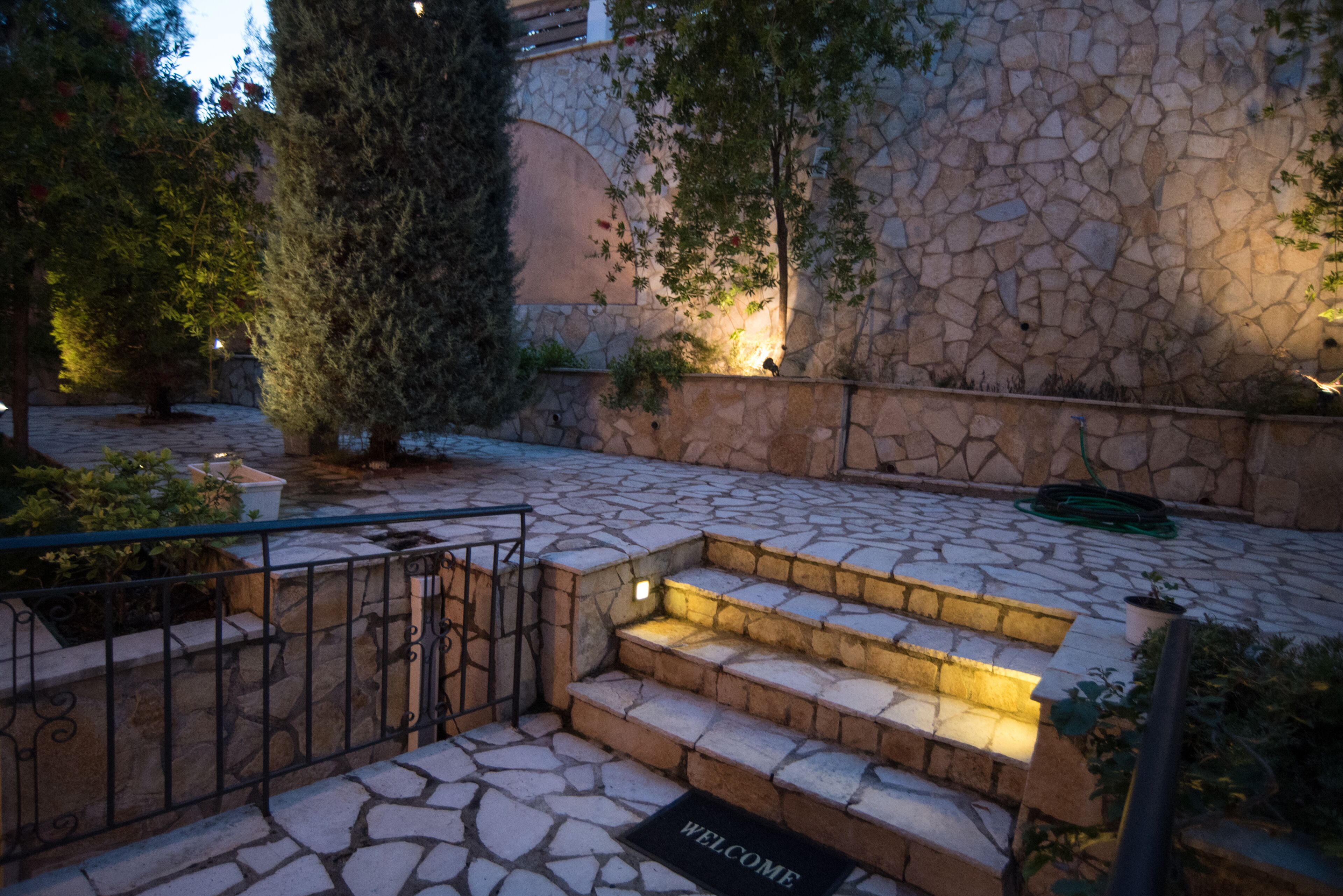 Una escalera de piedra conduce a una puerta con un felpudo que dice "WELCOME", rodeada de follaje y un muro de piedra.