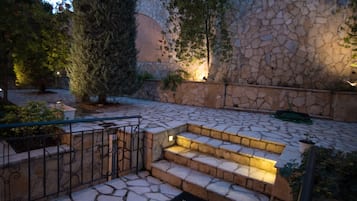 Una escalera de piedra conduce a una puerta con un felpudo que dice "WELCOME", rodeada de follaje y un muro de piedra.
