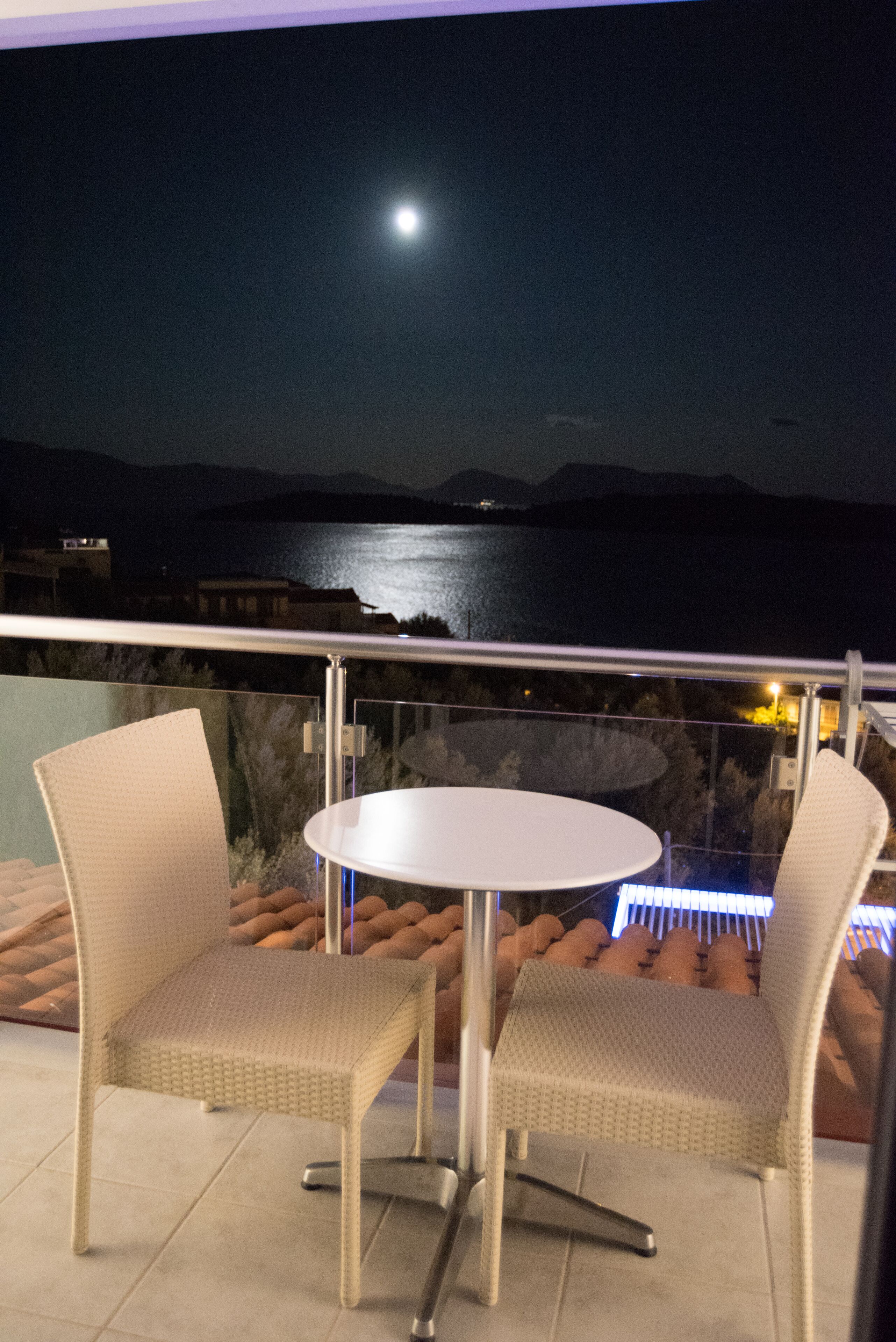 Un balcón con dos sillones de mimbre y una mesa redonda pequeña, con vista a una masa de agua y montañas distantes en la noche.