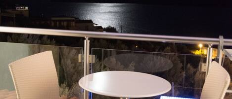 Un balcón con dos sillones de mimbre y una mesa redonda pequeña, con vista a una masa de agua y montañas distantes en la noche.