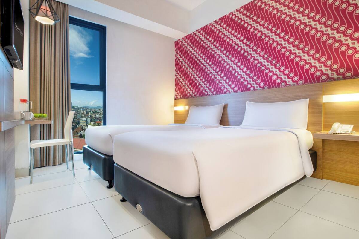 A modern hotel room with a large bed, a desk with a chair, a view of the city, and a patterned wall.